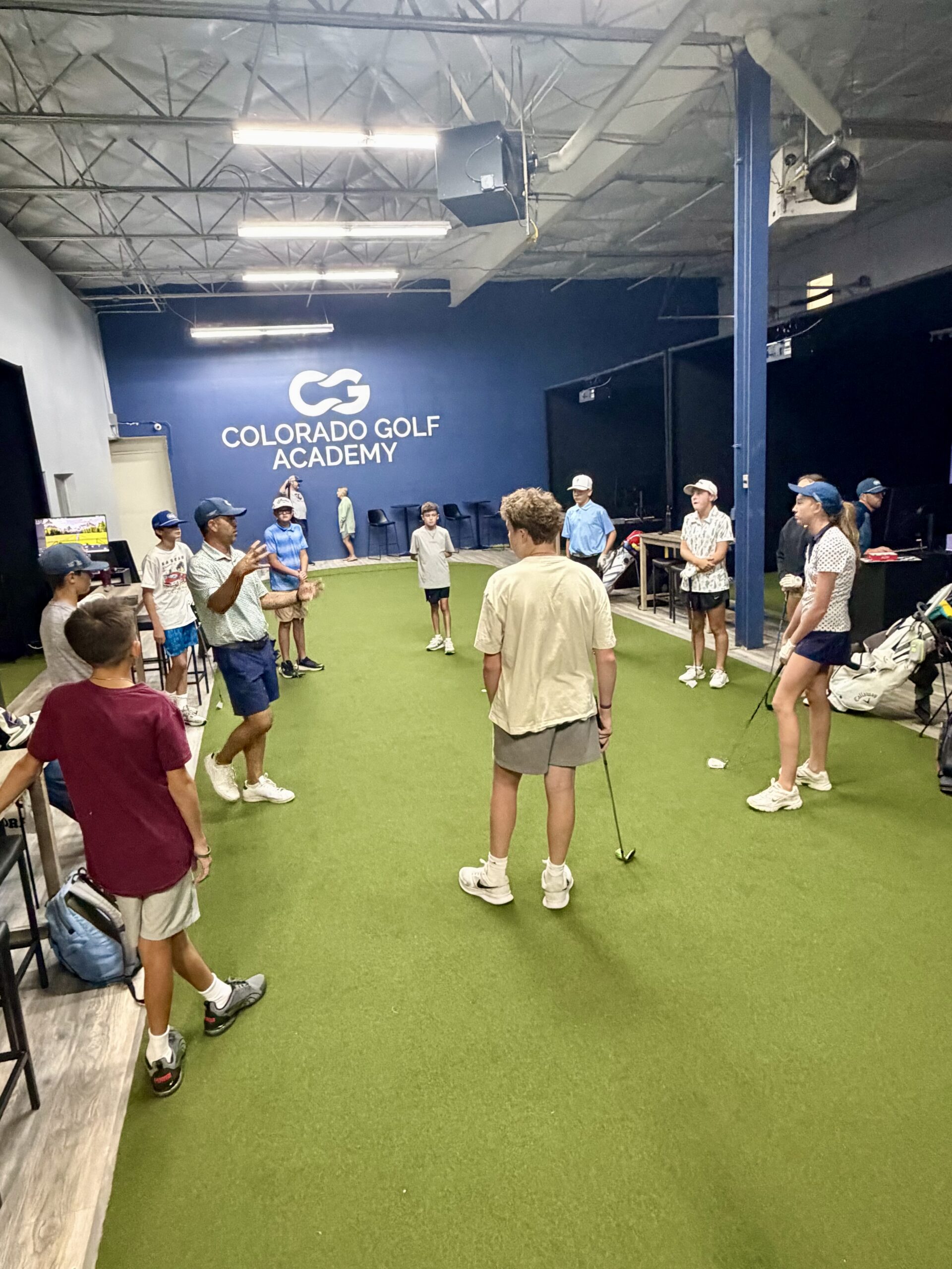 A group of boys standing in a loose circle around an instructor at an indoor golf course.